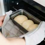 Homemade bread dough placed in oven with glove-covered hands. Fresh baking moment captured.